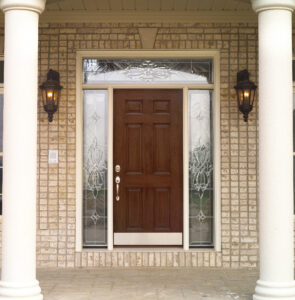 Lovely simulated redwood front door with decorative glass sidelights and overhead glass inset in brick cladding on front porch between two ivory-colored Greek-style columns 