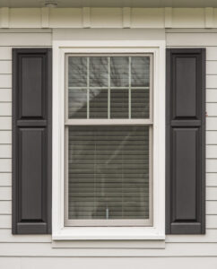 Exterior view of single-hung window with tan frame and grids in top sash, white casing, black shutters, on exterior wall with cream-colored siding