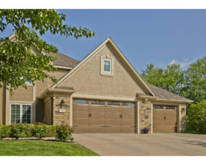 Upscale tan stucco house with stone accents and overhead carriage-style garage doors in a medium-brown color 