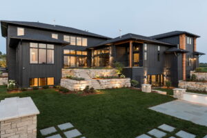 Rustic, luxury two-story home in modern design with gray lap siding, stone terraces, and multiple window styles, shown at twilight with interior lights glowing