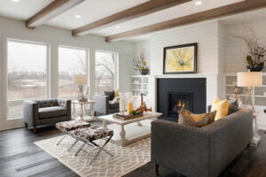 Modern living room with black fireplace, slate-colored sofa and chair, wood ceiling beams, and three large picture windows looking out on a wintry landscape