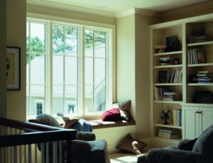 Interior shot of a set of three tall, slender windows with ivory frames and grids above a window seat in a loft room with bookshelves and easy chairs