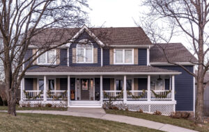 Two-story farmhouse-style home with dark blue siding, tan window shutters and white trim for the windows, porch columns and railings